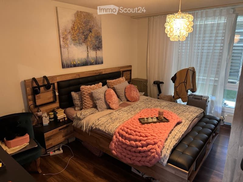 Cozy bedroom with a large bed, nightstand, chair, and a painting on the wall. The bed is adorned with multiple pillows and a pink throw blanket. A nightstand holds books and a lamp, and a stool is placed at the foot of the bed.