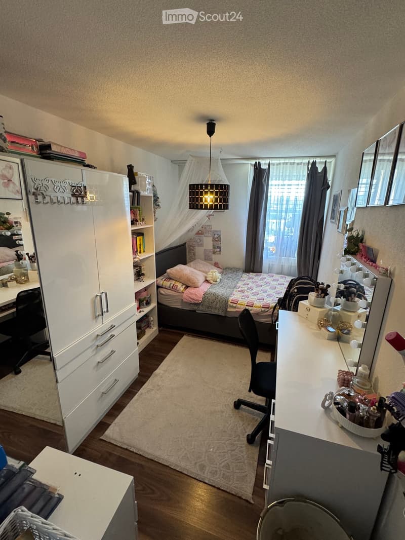 Cozy bedroom with a bed, desk, chair, and cabinet. Features a hanging lamp, a mirror, and a small rug on the floor.