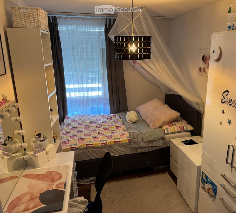 Cozy bedroom with a bed, colorful bedding, a hanging lamp, a desk with a mirror, and a chair. The room features a large window with curtains and a storage unit with a wicker basket on top.