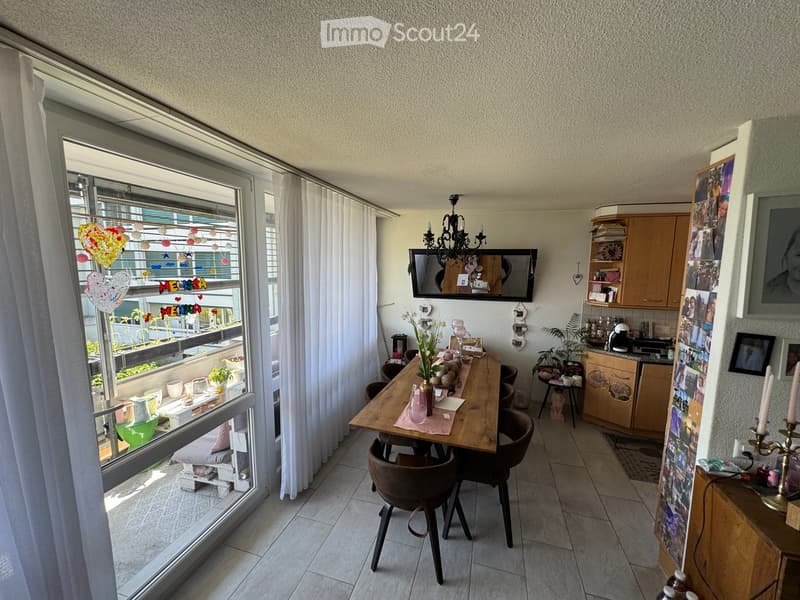 Bright living-dining area with large windows and balcony access, featuring a wooden dining table, chairs, cabinets, and a TV mounted on the wall.