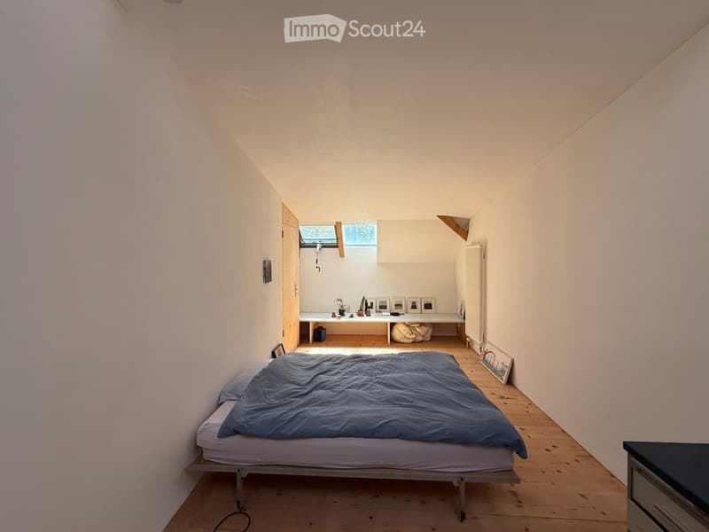 A compact bedroom with a single bed, nightstand, radiator, small table with chairs, and picture frames on the wall. The room features wooden flooring and white walls, with a skylight providing natural light.