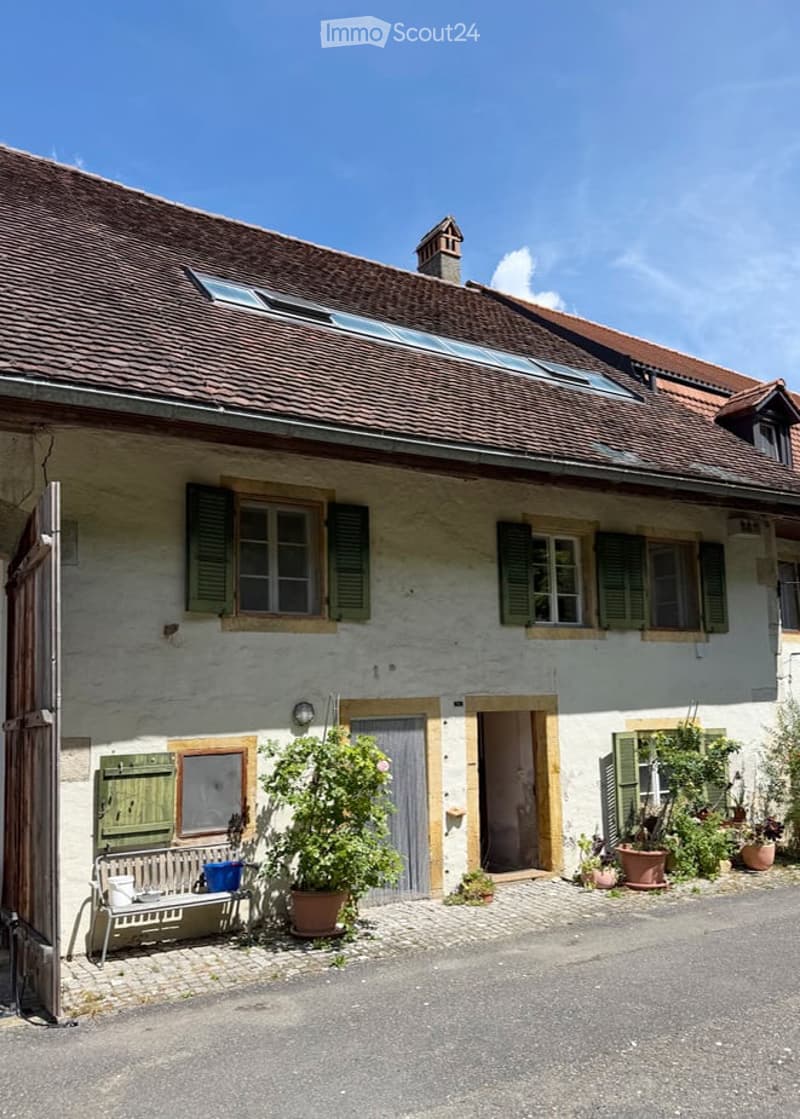 A charming, rustic house with a tiled roof, white exterior, and green shutters. The entrance features a wooden door, a small bench, and potted plants, creating a welcoming atmosphere.