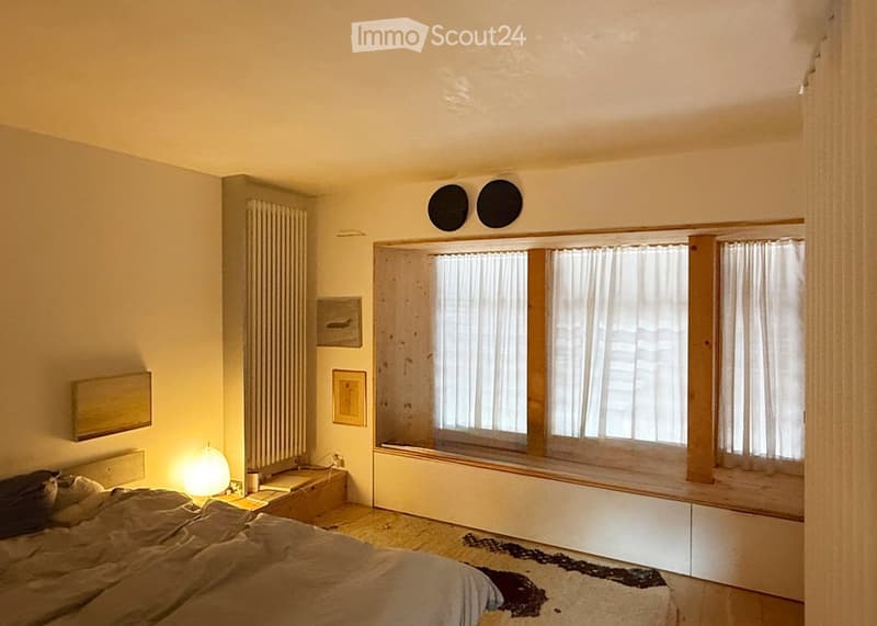 Bright bedroom with wooden floor, large window with white curtains, bed with pillows and blanket, nightstand with lamp, picture frames on wall, radiator, and a small rug on the floor.