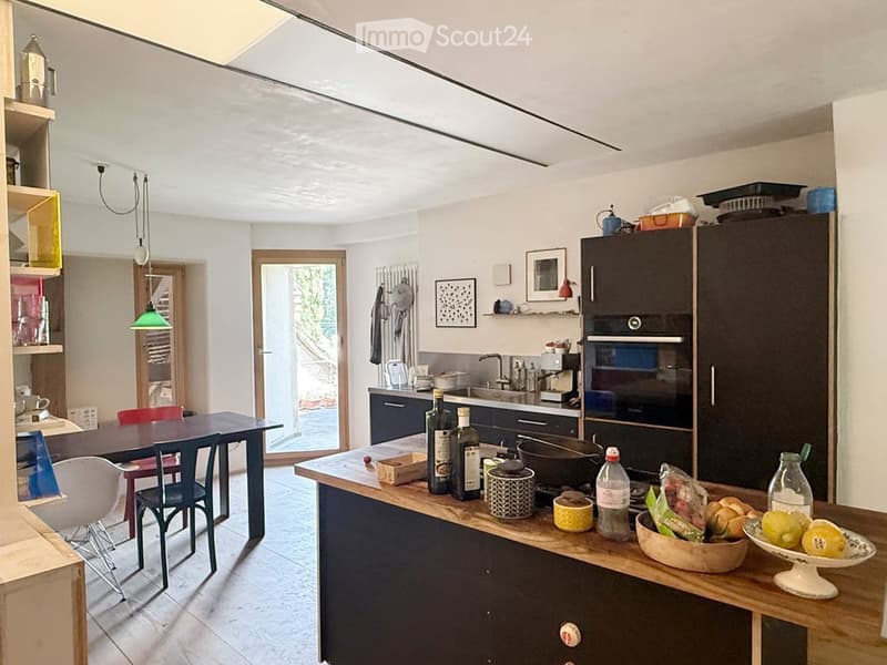 Modern kitchen with dark cabinets, wooden island, and various appliances. Includes a dining table with chairs, a bowl of fruit, and several bottles on the counter.
