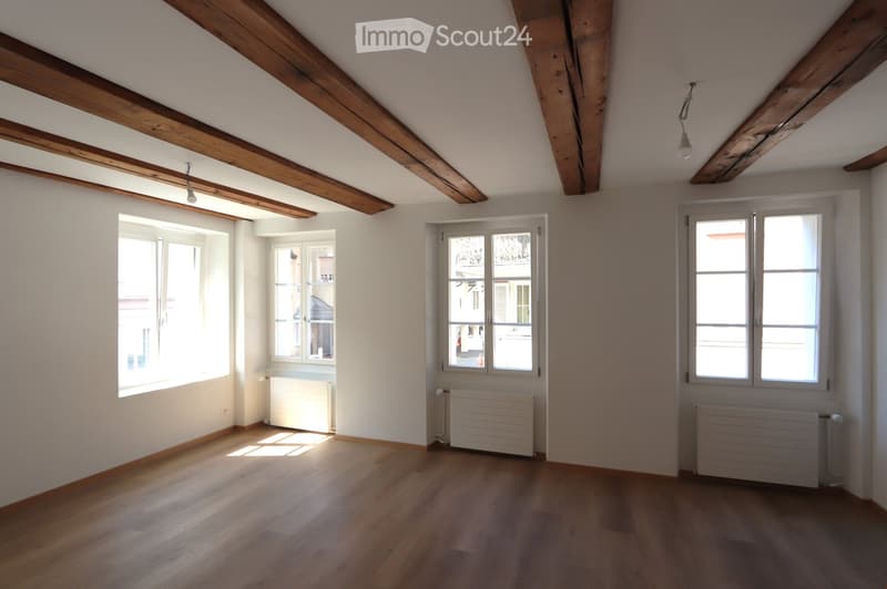 Empty attic room with wooden beams, wooden flooring, and multiple windows allowing natural light. Radiators are present along the walls.