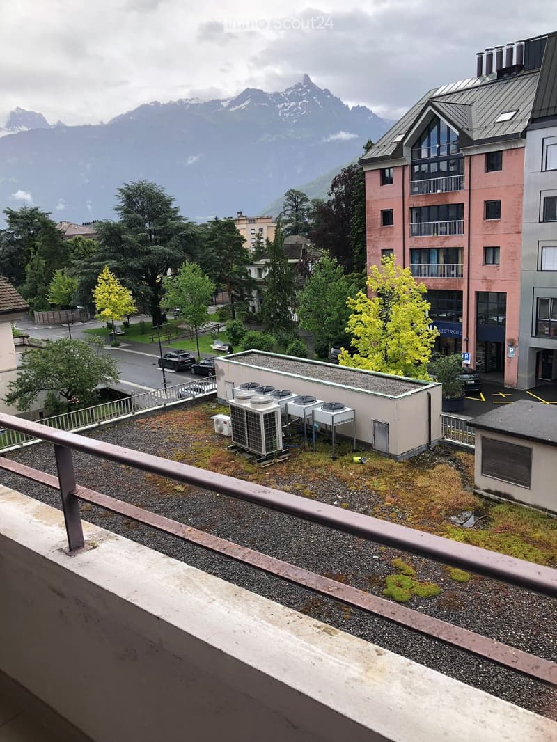 View of a multi-story residential building with balconies, surrounded by trees and a mountainous backdrop. Rooftop area visible with air conditioning units and gravel surface.