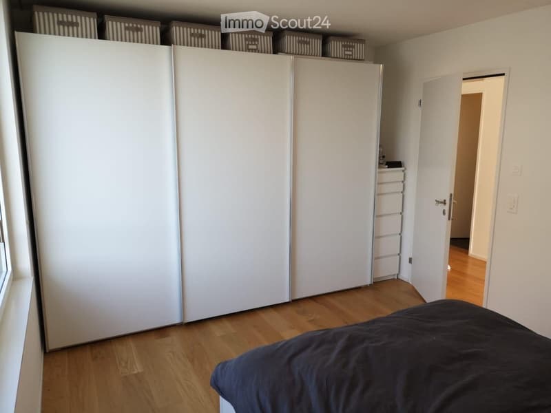 Modern bedroom with wooden flooring, large white wardrobe, and a neatly made bed with dark bedding.