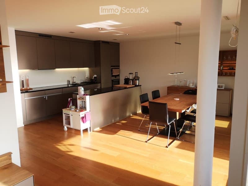 Modern kitchen with grey cabinetry, stainless steel appliances, a central island, and a dining area with a wooden table and chairs. The space features wooden flooring and ample natural light from a skylight.