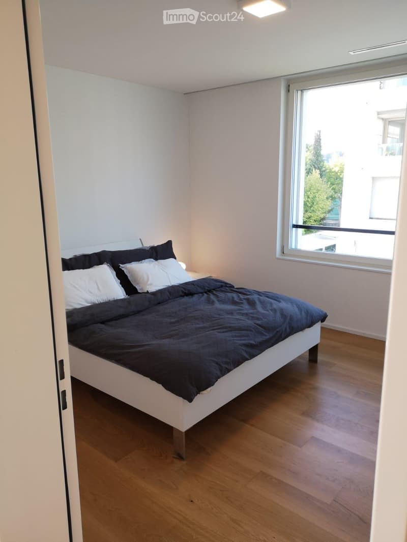 Modern bedroom with a white bed, dark blue blanket, and pillows, set on a wooden floor with a large window offering a view of greenery.
