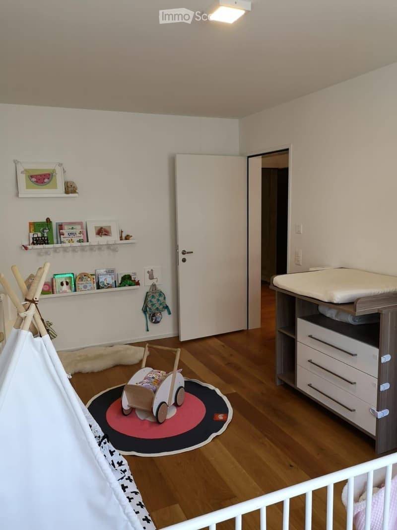 Brightly lit children's bedroom with wooden flooring, a crib, changing table, toy teepee, and shelves with books and toys.