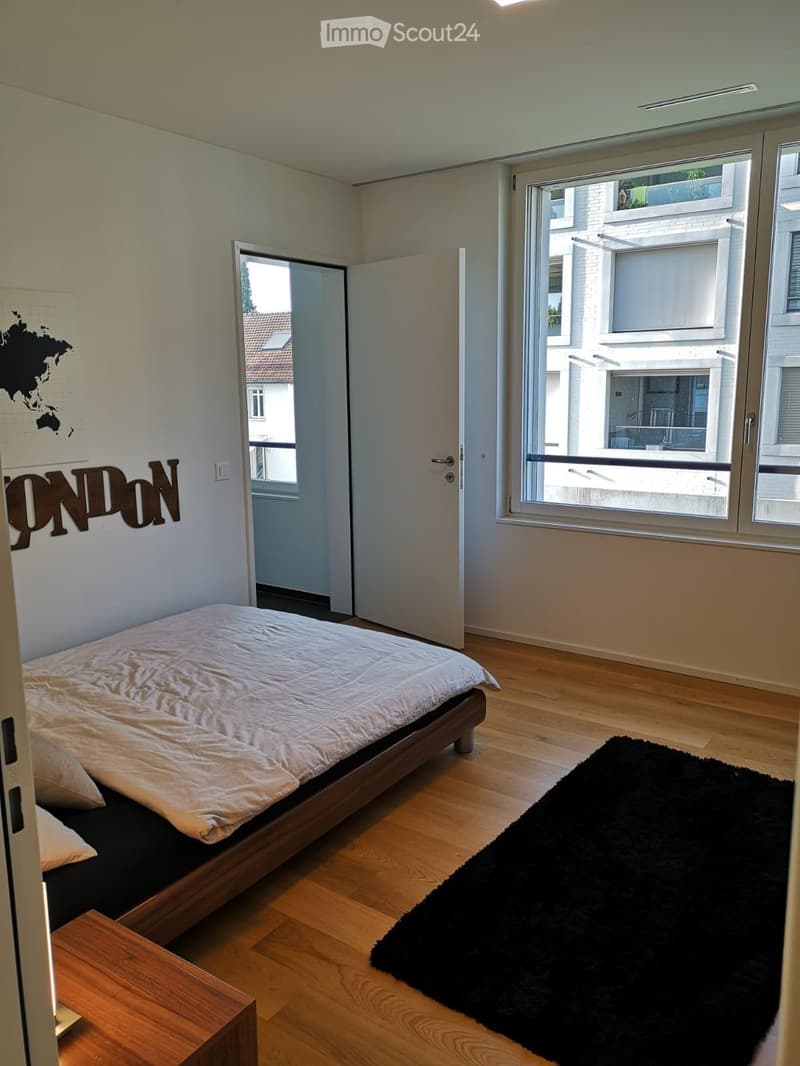 Modern bedroom with wooden flooring, a bed with white bedding, a black rug, and a nightstand. The room features large windows with a view of the city and a door leading to a balcony.