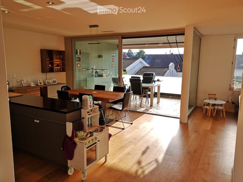 Bright, open-plan living and dining area with wooden flooring, large glass doors leading to a balcony, dining table with chairs, kitchen area with cabinets and appliances, and a small play kitchen for children.