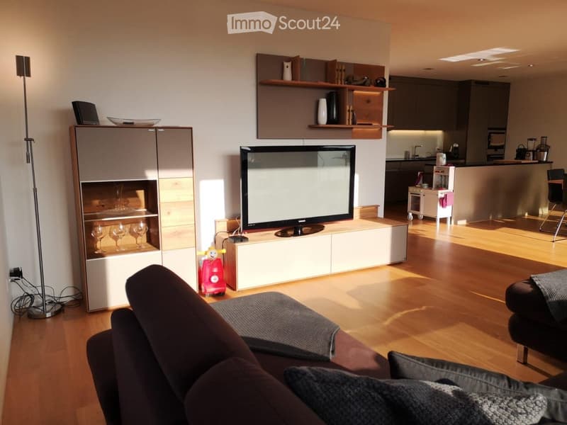 Modern living room with a dark brown sofa, ottoman, flat-screen TV on a white stand, glass cabinet with wine glasses, floor lamp, and an open kitchen area with a counter and chairs.