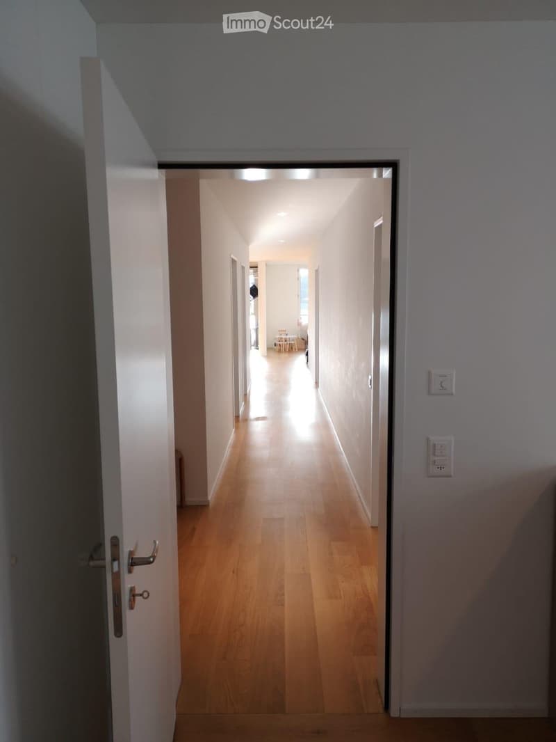 A narrow hallway with light wooden flooring and white walls, leading to a room with a small table and chairs visible at the end.