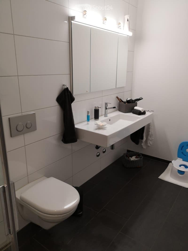 Modern bathroom with white tiles, black floor, sink, toilet, mirror, and various accessories including a wicker basket and a small toilet seat for children.