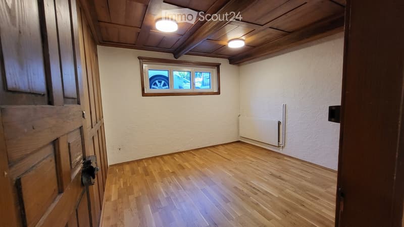 Empty room with wooden flooring, white walls, and a window showing a car outside. The room has exposed wooden beams on the ceiling and a radiator on one wall.
