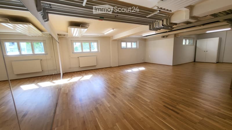 Empty room with wooden flooring, white walls, exposed ceiling pipes, and several windows allowing natural light.