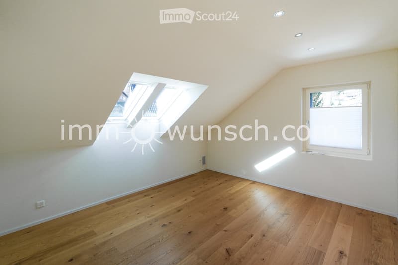 Empty attic room with wooden flooring, white walls, and a skylight window, featuring recessed ceiling lights and a regular window with blinds.