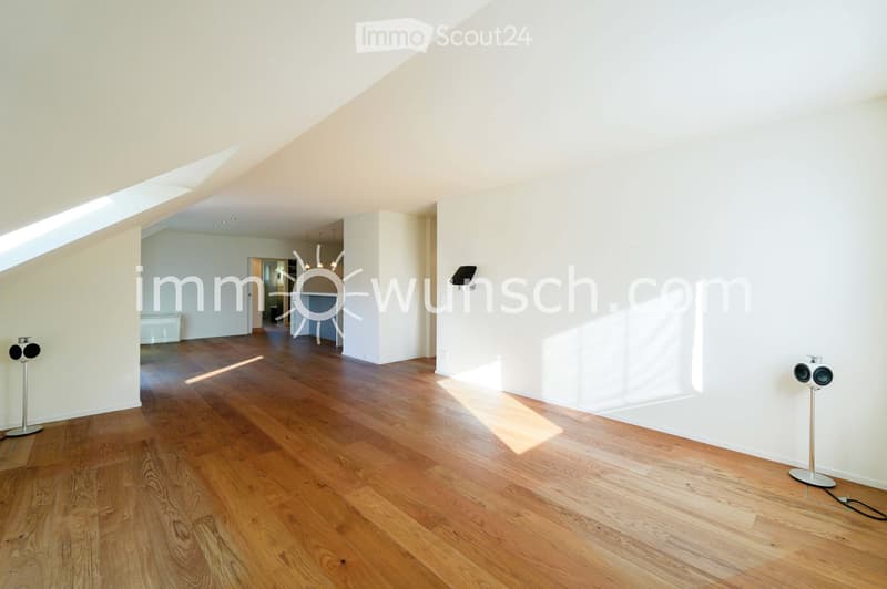 Bright, empty room with wooden flooring and white walls, featuring large windows and a skylight allowing natural light to fill the space.