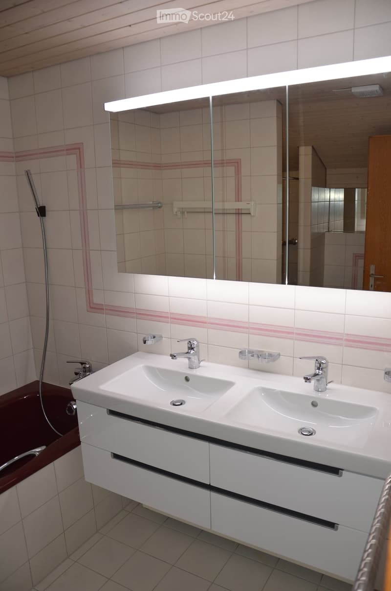 Modern bathroom with a double sink, large mirror, bathtub, and shower hose. The tiles are white with pink accents, and the sink has a sleek, contemporary design with modern faucets.
