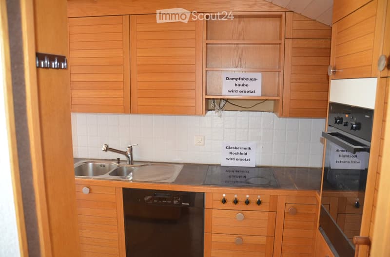 Modern kitchen with wooden cabinets, stainless steel sink, faucet, oven, and dishwasher. Signs indicate upcoming replacements for the range hood and glass ceramic cooktop.