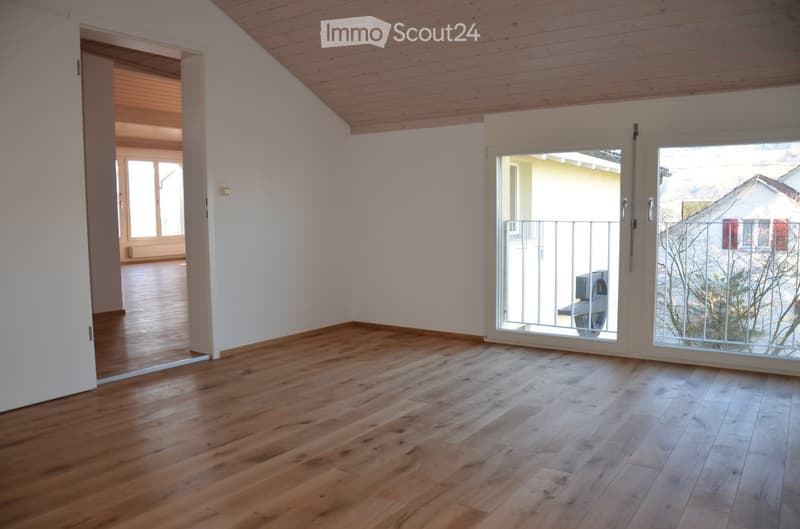 Empty room with wooden flooring, white walls, and large windows leading to a balcony area. The ceiling is slanted with wooden paneling.