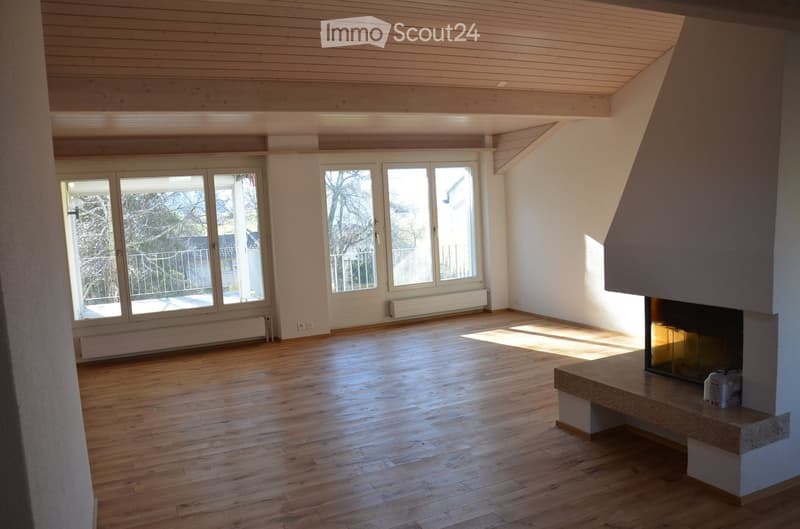 Bright, empty room with wooden flooring, large windows, and a fireplace with a stone surround. The ceiling has exposed wooden beams.