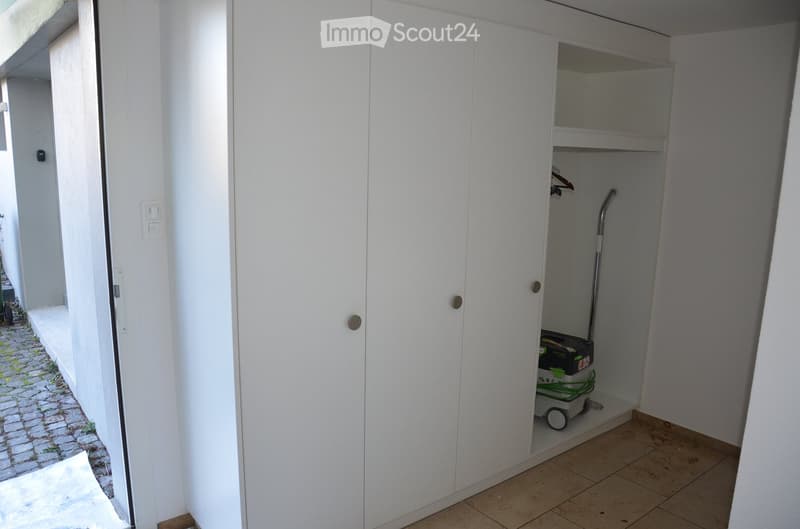 A small, white-walled closet with a vacuum cleaner stored inside, featuring a tiled floor and a partially open door leading to an outdoor area.