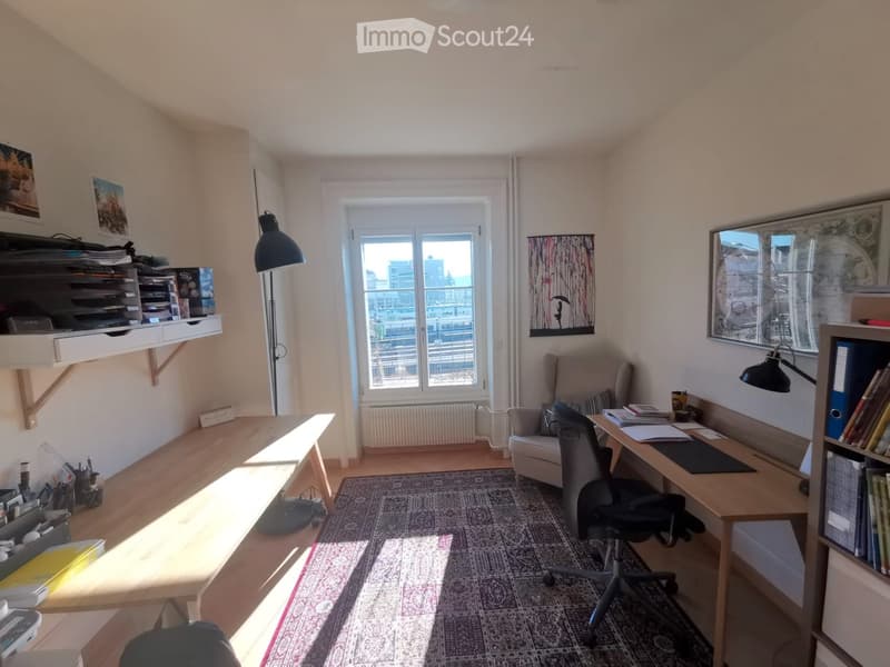 Brightly lit room with wooden floors, large window, desk, chair, sofa, bookshelf, and artwork on walls.