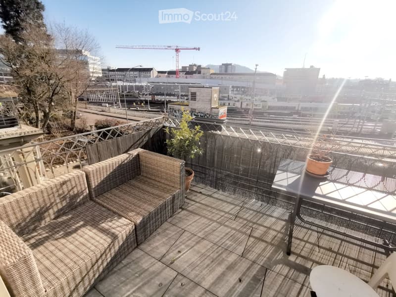 Balcony with a wicker sofa, glass table, and potted plants, overlooking a cityscape with a train station and construction cranes.