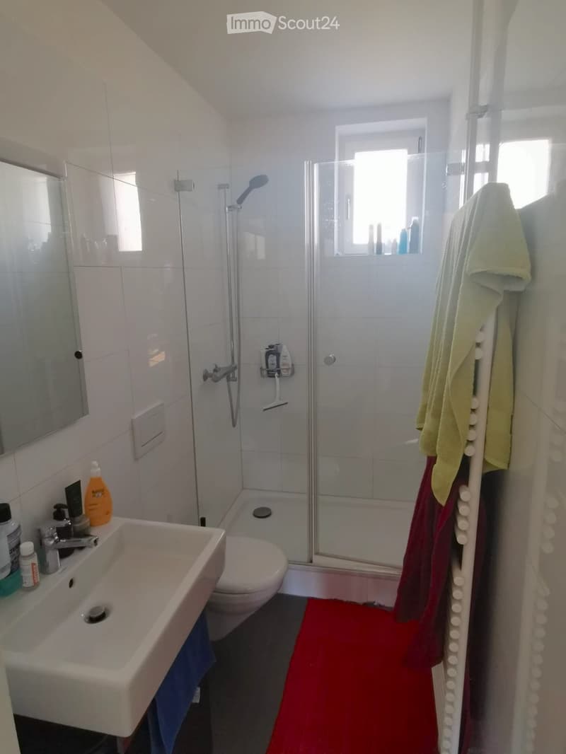Modern bathroom with white tiles, a sink, toilet, shower, and a towel rack with towels. The shower has a glass enclosure and a window above it.