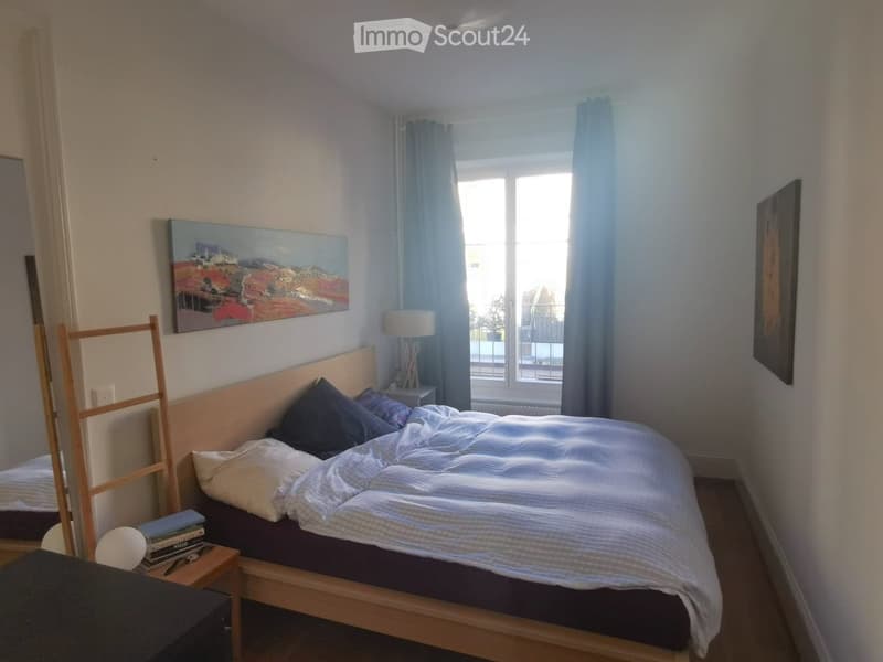 Cozy bedroom with a double bed, nightstand, lamp, and artwork on the walls. The room features a window with curtains and a small balcony view.