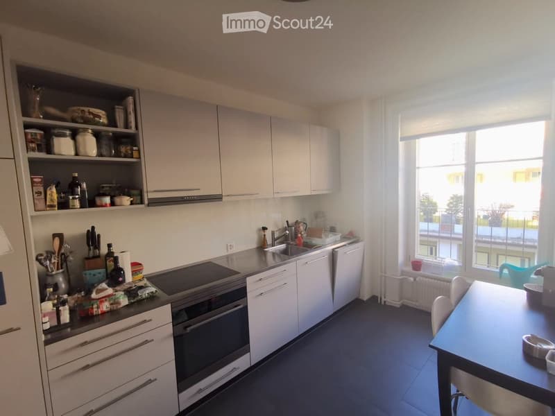 Modern kitchen with white cabinets, stainless steel appliances, and a large window overlooking a balcony. The counter is cluttered with various kitchen items, including utensils, bottles, and food packages.