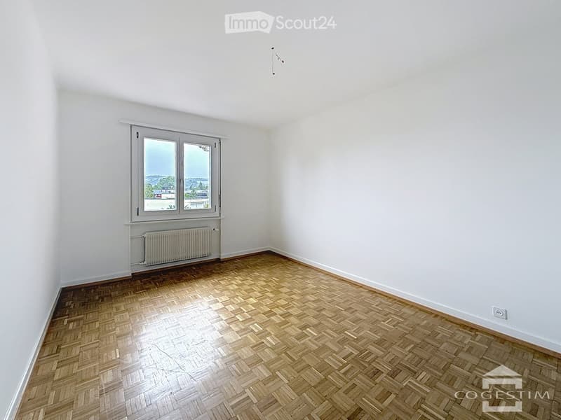 Empty room with wooden herringbone flooring, white walls, and a large window with a radiator below it.