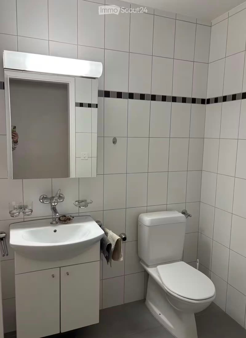 Modern bathroom with white tiles, a white sink with a chrome faucet, a mirrored cabinet, and a toilet. A towel hangs on a rack next to the sink.
