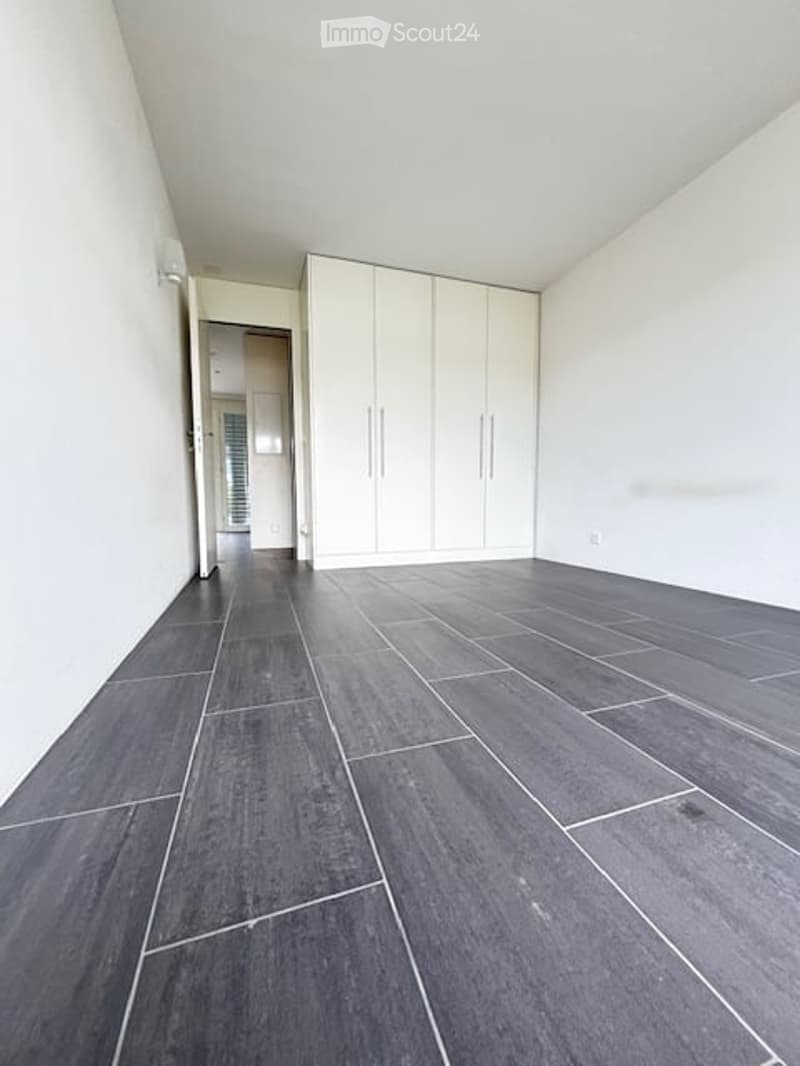 Empty room with dark grey tiled flooring and white walls. A white built-in wardrobe is visible along one wall, and an open doorway leads to another room.