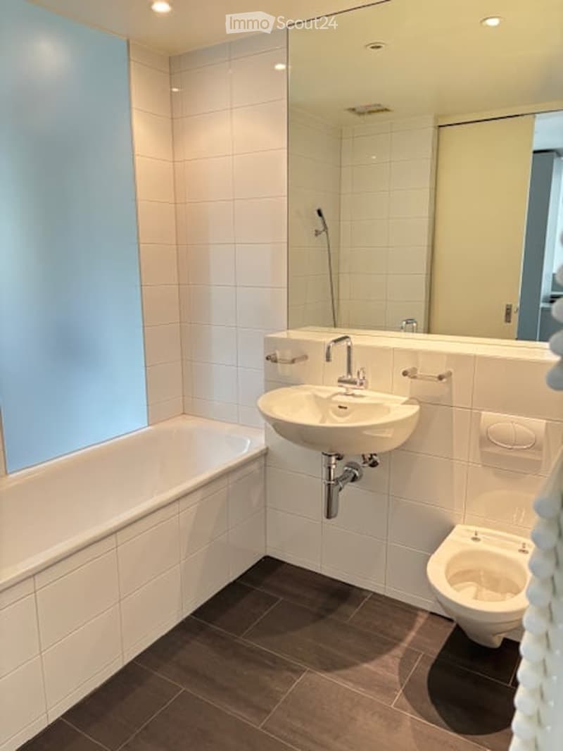 Modern bathroom with white tiles, a bathtub, a sink with a chrome faucet, a toilet, and a large mirror above the sink.