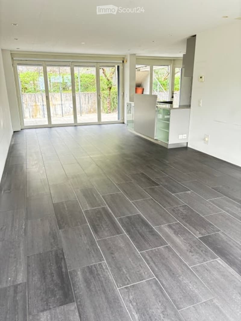 Open-plan space with dark grey tiled flooring, large glass sliding doors leading to an outdoor area, and a small white kitchen unit visible in the corner.