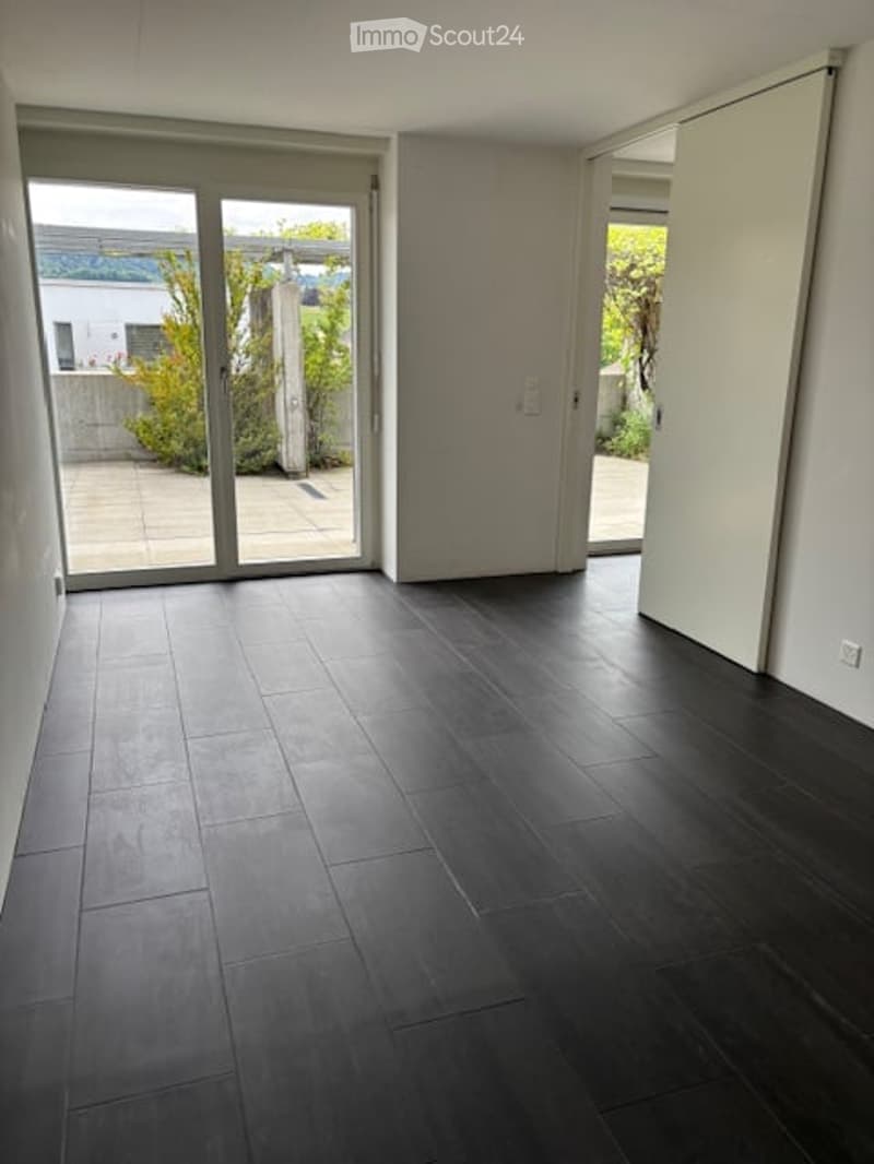 Completely empty room with dark tiled flooring, large glass sliding doors leading to an outdoor area, and white walls.
