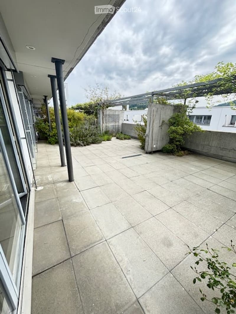 Spacious balcony with concrete tiles, modern railings, and a view of greenery and neighboring buildings.
