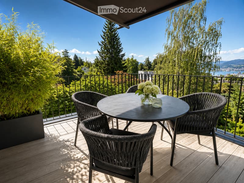 Balcony with a round table, four wicker chairs, and a vase of white flowers, overlooking a scenic view of trees and a body of water.