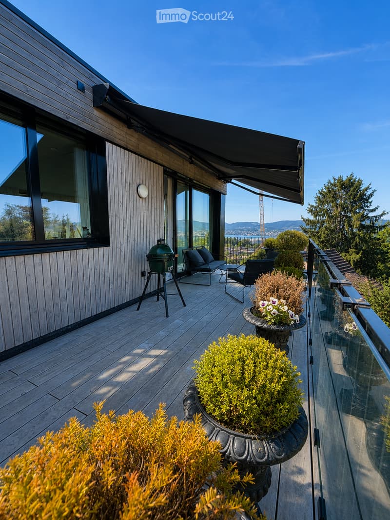 Wooden balcony with modern furniture, grill, and scenic view of mountains and water.