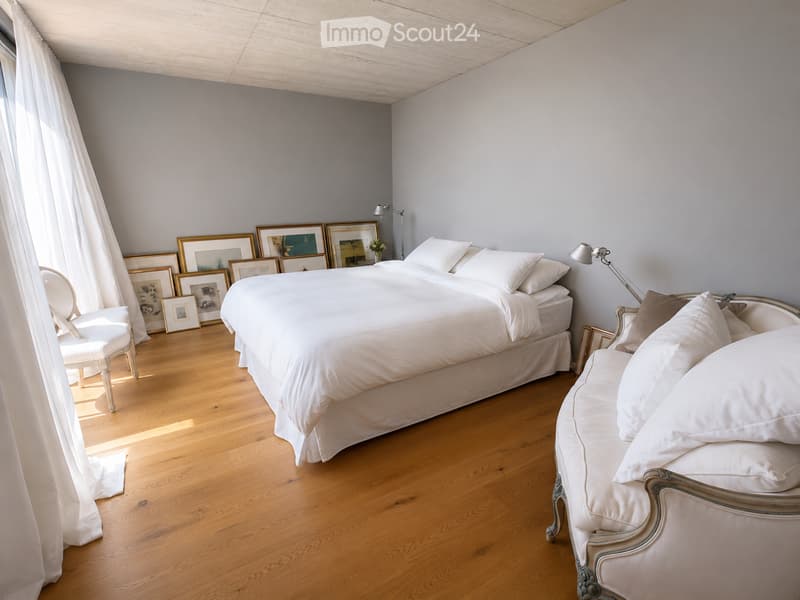 Bright bedroom with wooden flooring, large bed with white bedding, sofa, chair, and several picture frames on the wall.