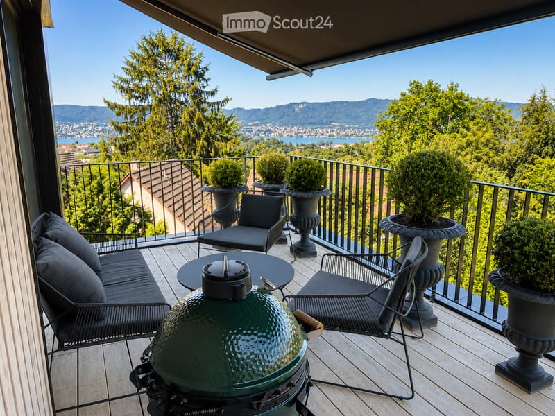 Balcony with wooden decking, black metal railing, and scenic mountain and lake view. Features a green grill, sofa, chairs, and potted plants.