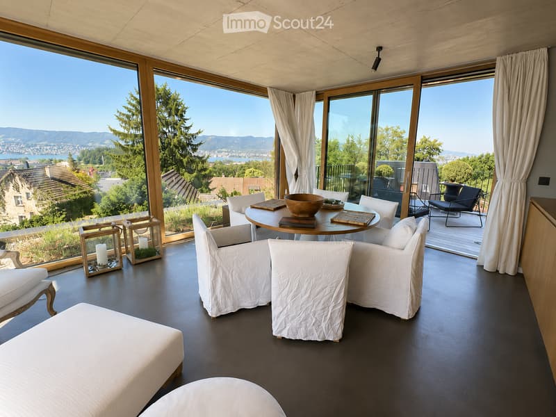 Modern living room with large windows offering scenic views, featuring white furniture including sofas, chairs, a round table, and decorative candle holders.