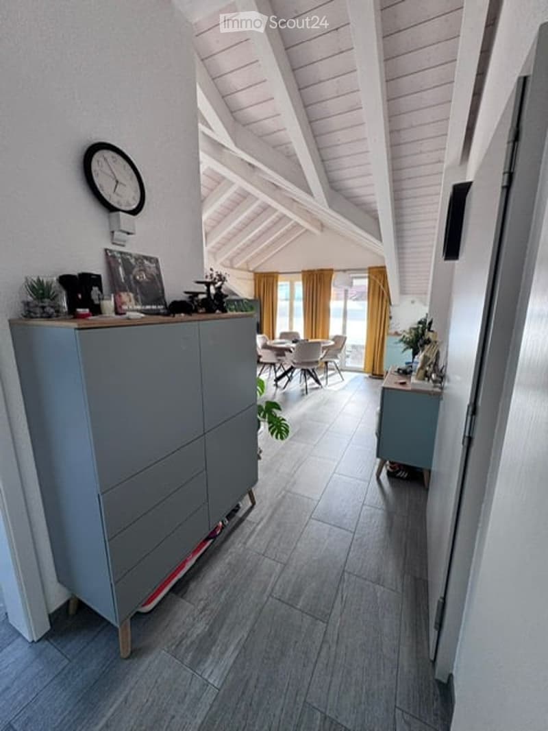 Modern hallway with grey cabinet, clock, picture frame, and a view into a dining area with a table and chairs. Wooden flooring and white walls.