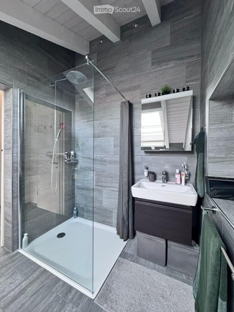 Modern bathroom with a glass-enclosed shower, a white sink with a dark wooden cabinet, and a mirror above the sink. The walls and floor are covered in grey tiles, and a towel hangs on the side.