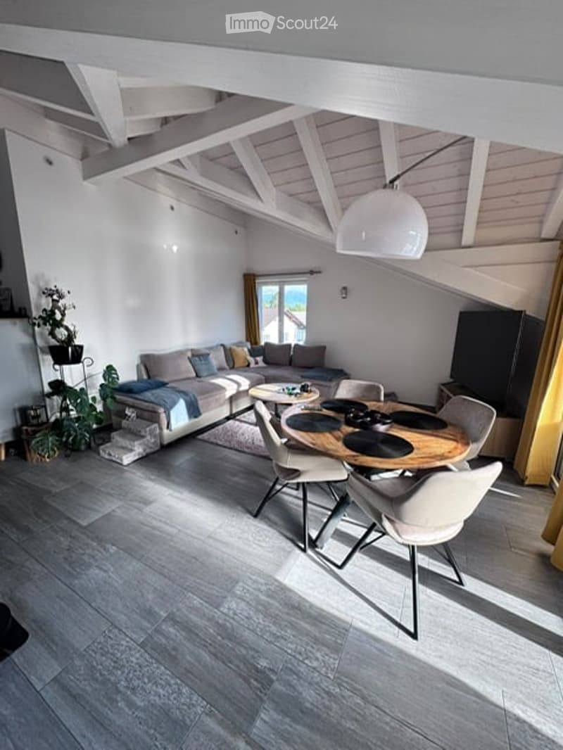 Bright, open-concept living and dining area with exposed wooden beams, grey sofa, coffee table, dining table with chairs, pendant light, and decorative plants.