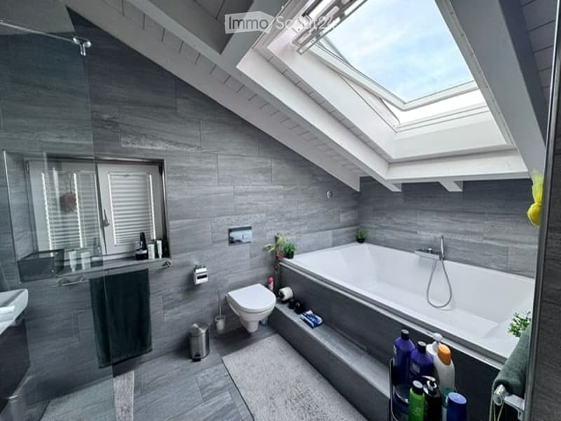 Modern bathroom with grey tiles, a large bathtub, toilet, sink, and a skylight window above the tub.