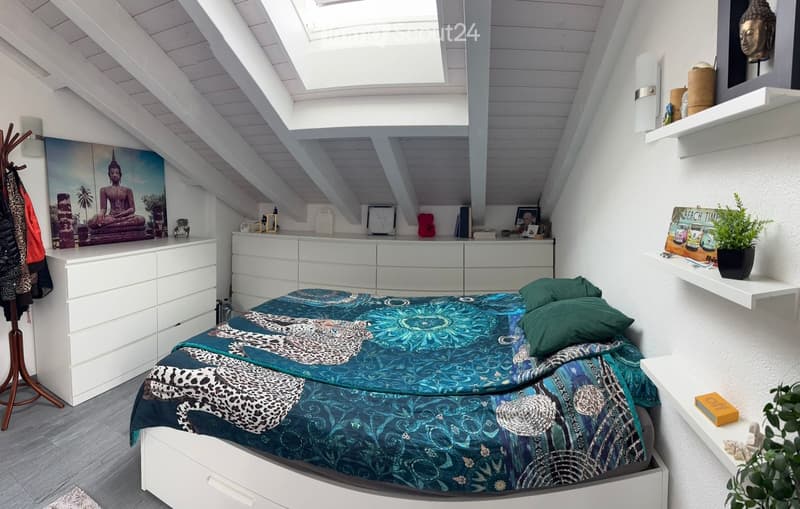 A modern bedroom with a slanted wooden ceiling, skylight, and white walls. Features a bed with a blue and black patterned duvet, green pillows, a white dresser, shelves with decorative items, and a coat rack with jackets.